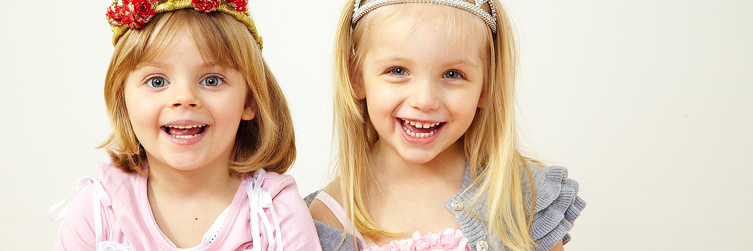 Zwei kleine blonde Mädchen sind als Prinzessinnen verkleidet, sitzen nebeneinander und lachen.